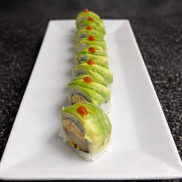 A row of avocado-topped sushi rolls with red dots on a white rectangular plate.